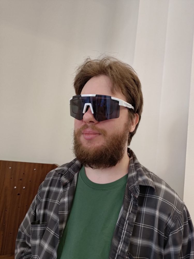 A man with short light-brown hair and a short-medium beard, wearing a grey chekered shirt over a green t-shirt, and giant racing-style sunglasses over regular eyeglasses.