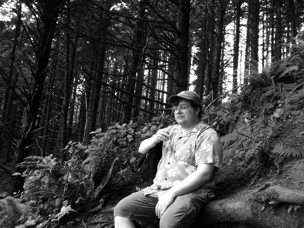 A man in a baseball cap, big glasses, a flower print shirt, and shorts sitting on a log in a forest drinking something from the cap of a thermos. His backpack is sitting next to him. High contrast monochrome digital photo.