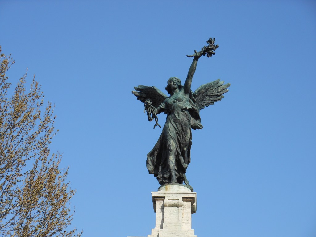 A green statue against a bright blue sky. The statue is of an angelic figure with a broken shackle held at about shoulder height and a plant of some sort held out above its head. It stands a marble plinth. There's a bit of a tree with orange/brown leaves poking in from the left side of the frame.